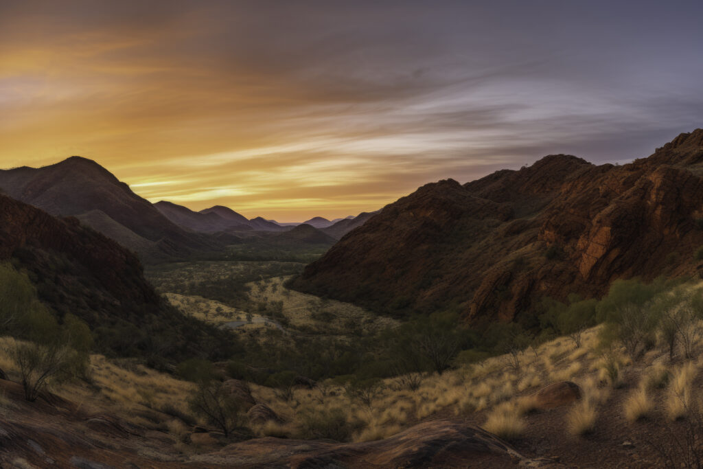 Delve into the striking beauty of the MacDonnell Ranges with this panoramic depiction. The image presents a comprehensive view of the landscape, revealing an intricate tapestry woven from rugged mountains, expansive valleys, and unspoiled wilderness. It's a captivating celebration of the outdoors where the natural splendor of the Australian outback unfolds before your eyes. This isn't just a view—it's an immersive journey into the heart of nature's grandeur.