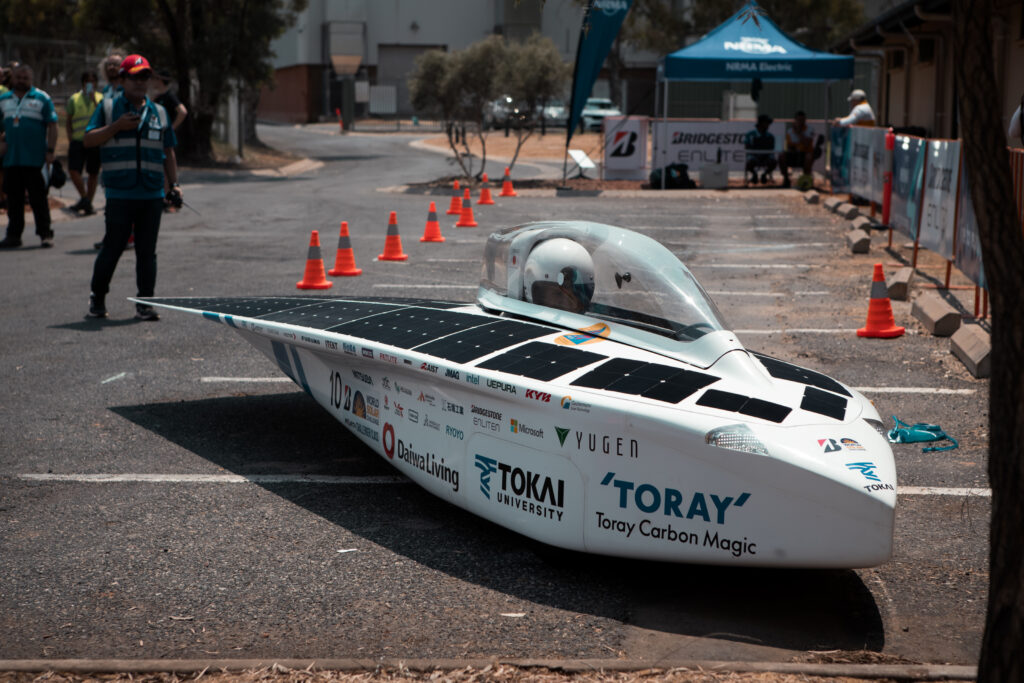 A vivid capture of the Bridgestone World Solar Challenge 2023. The image is centred around an enthusiastic participant, ready for the challenge at www.worldsolarchallenge.org. He is accessorised with essentials such as a helmet, gloves, and sturdy shoes. Key highlights include his eco-friendly boat vehicle, carved ingeniously from Toray Carbon Magic, symbolising the collaboration of University talent with industry giants like NUMA, BRIDGESTO, ENLITE, YUGEN, and TOKAI. The scene is set by a backdrop of sophisticated cars, rounding out a vibrant visual narrative about the confluence of technology, passion, and sustainability.